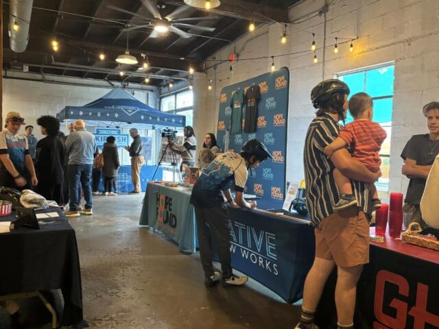 Indoor community event with multiple info booths, string lights, and people browsing tables along the room.