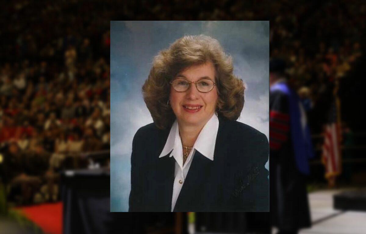 Smiling middle-aged woman with curly hair and glasses in a navy blazer, studio portrait.