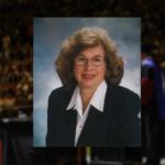 Smiling middle-aged woman with curly hair and glasses in a navy blazer, studio portrait.