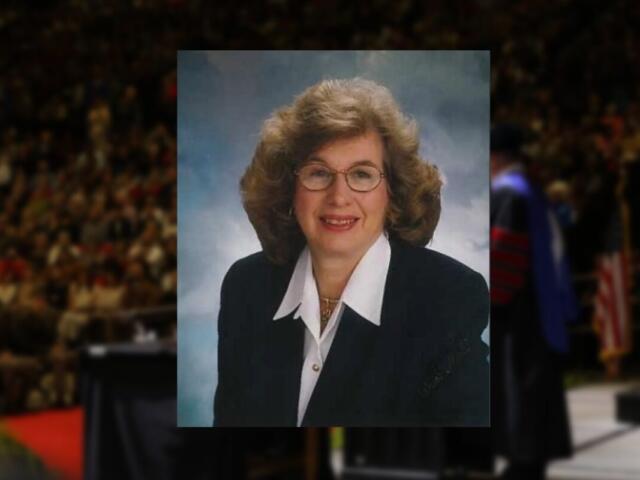 Smiling middle-aged woman with curly hair and glasses in a navy blazer, studio portrait.