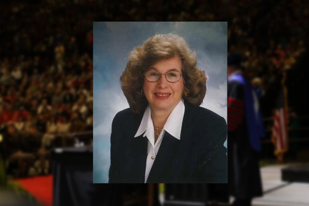 Smiling middle-aged woman with curly hair and glasses in a navy blazer, studio portrait.