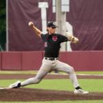Baseball pitcher in black uniform throwing a pitch from the mound, left leg extended forward and body in motion.