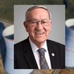 Portrait of an older man in a dark suit and tie, smiling at the camera; blurred industrial background behind him.
