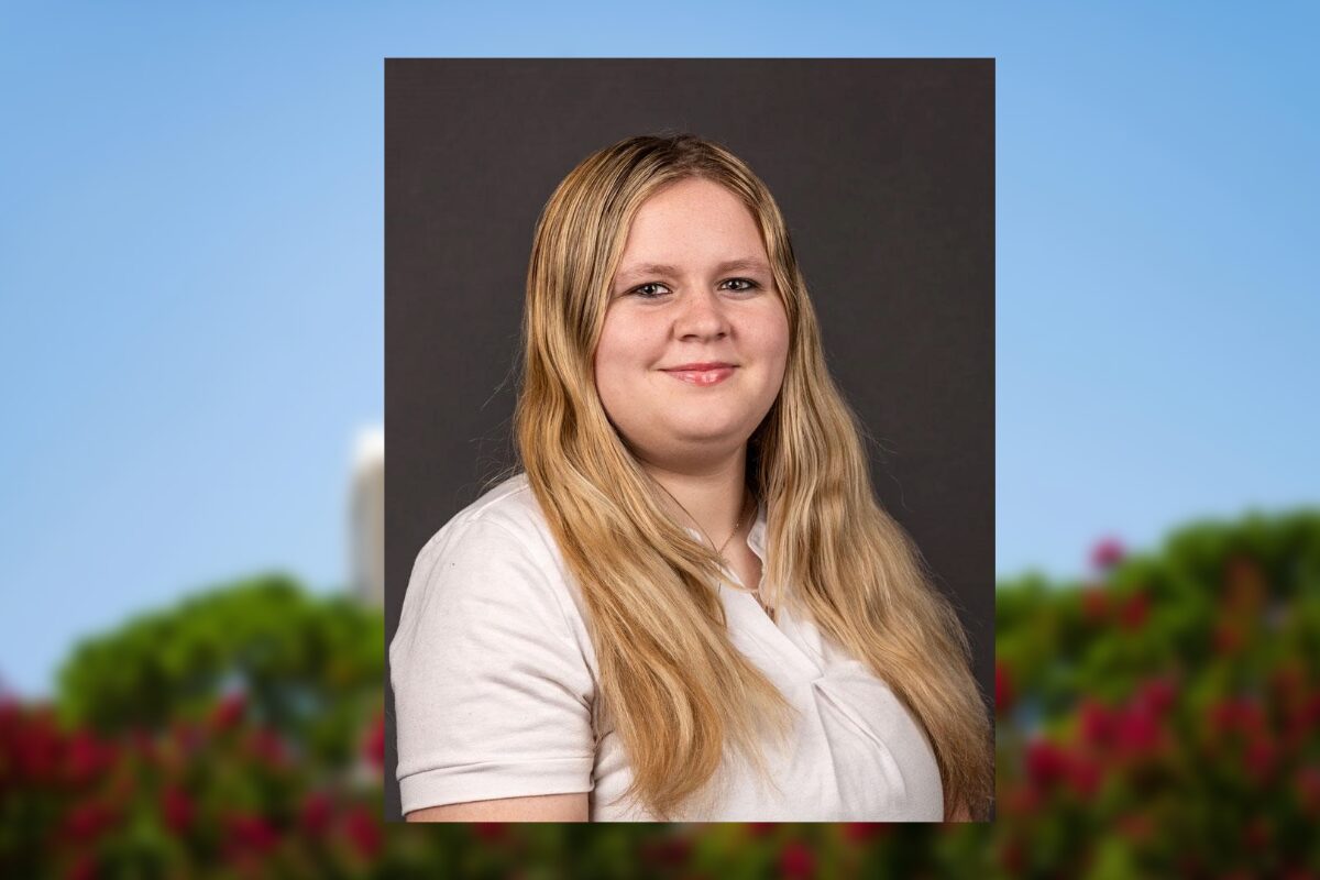 Portrait of a smiling woman with long blonde hair, wearing a white blouse, against a dark gray backdrop.