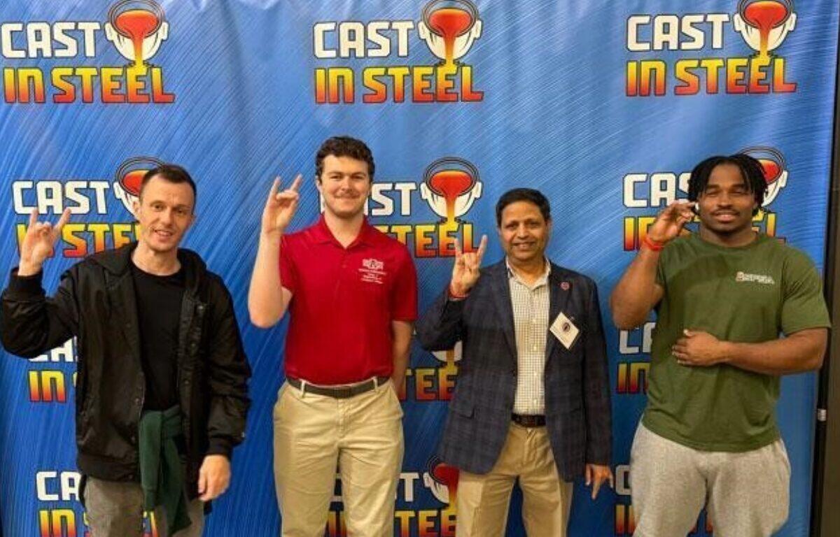 Four men stand in front of a blue Cast in Steel backdrop, each making the rock-on hand gesture and smiling.