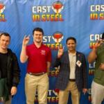 Four men stand in front of a blue Cast in Steel backdrop, each making the rock-on hand gesture and smiling.