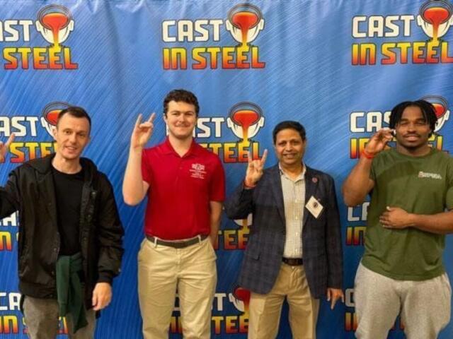 Four men stand in front of a blue Cast in Steel backdrop, each making the rock-on hand gesture and smiling.