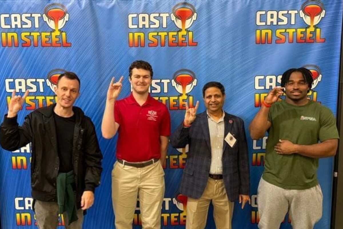 Four men stand in front of a blue Cast in Steel backdrop, each making the rock-on hand gesture and smiling.