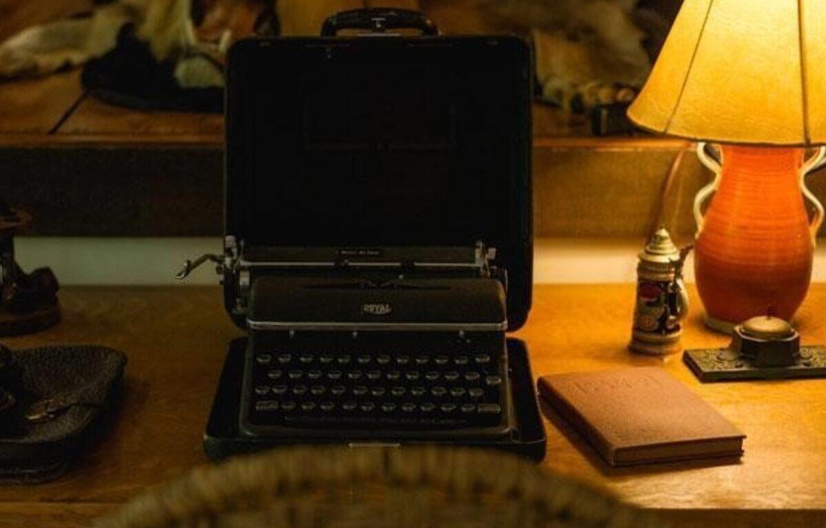 Vintage portable typewriter in an open case sits on a wooden desk beside a warm lamp and a notebook on the right.