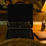 Vintage portable typewriter in an open case sits on a wooden desk beside a warm lamp and a notebook on the right.