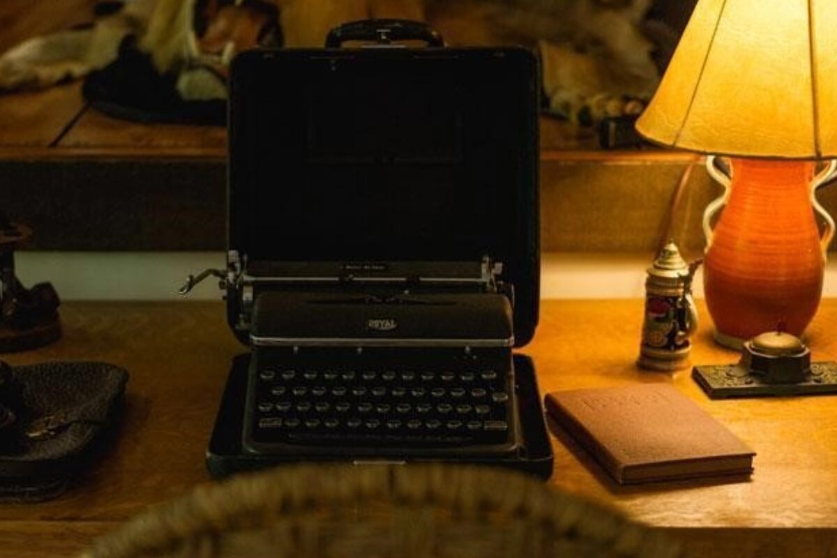 Vintage portable typewriter in an open case sits on a wooden desk beside a warm lamp and a notebook on the right.