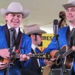 Three musicians in matching blue suits and white cowboy hats perform on a stage; two play guitars and one plays a mandolin with microphones nearby.