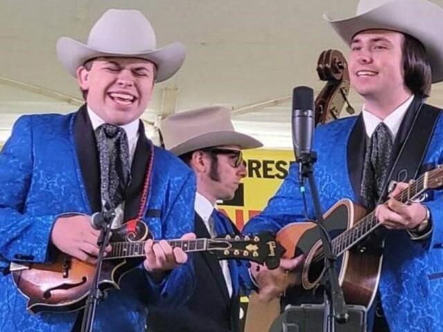 Three musicians in matching blue suits and white cowboy hats perform on a stage; two play guitars and one plays a mandolin with microphones nearby.