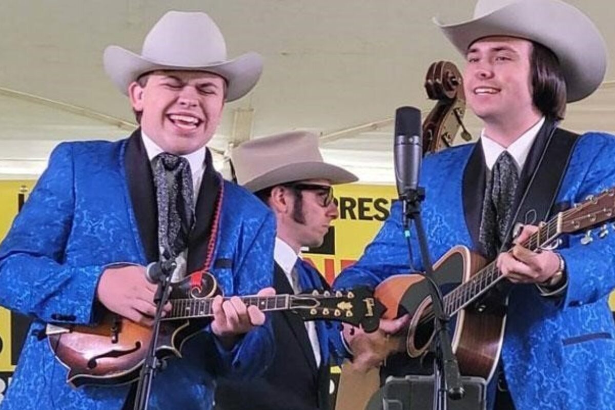 Three musicians in matching blue suits and white cowboy hats perform on a stage; two play guitars and one plays a mandolin with microphones nearby.