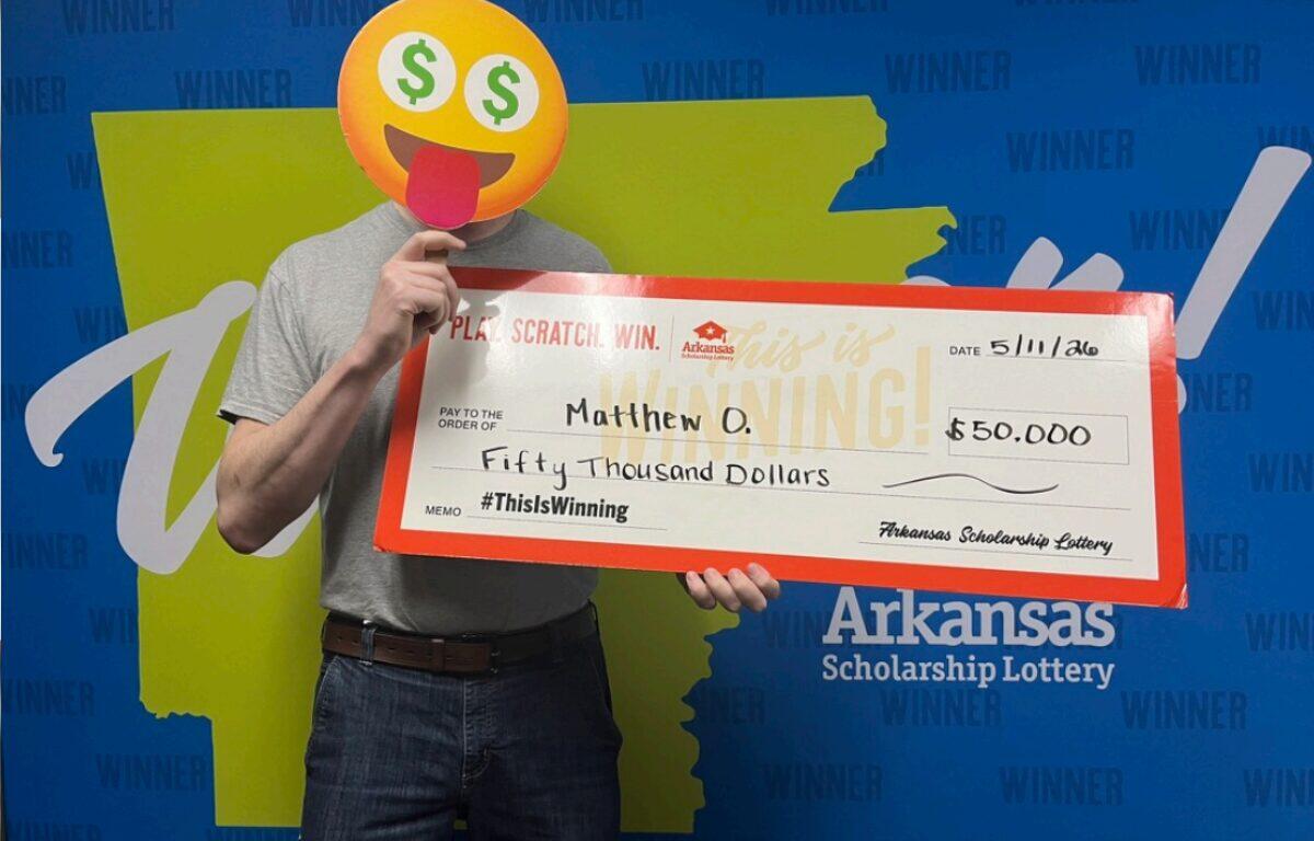 Person with an emoji face mask holds a giant Arkansas Scholarship Lottery check for $50,000, standing in front of a 'WINNER' backdrop.