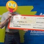 Person with an emoji face mask holds a giant Arkansas Scholarship Lottery check for $50,000, standing in front of a 'WINNER' backdrop.
