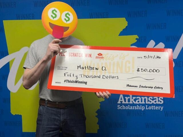 Person with an emoji face mask holds a giant Arkansas Scholarship Lottery check for $50,000, standing in front of a 'WINNER' backdrop.