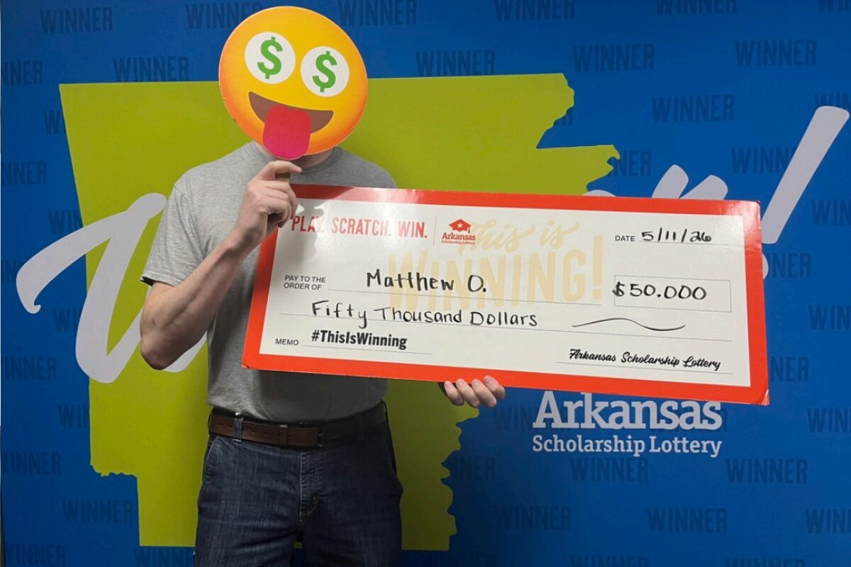 Person with an emoji face mask holds a giant Arkansas Scholarship Lottery check for $50,000, standing in front of a 'WINNER' backdrop.