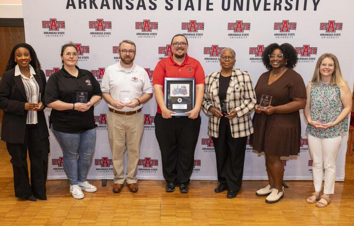 Group of seven people standing in front of an Arkansas State University backdrop, each holding an award or plaque at an event.
