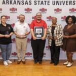 Group of seven people standing in front of an Arkansas State University backdrop, each holding an award or plaque at an event.