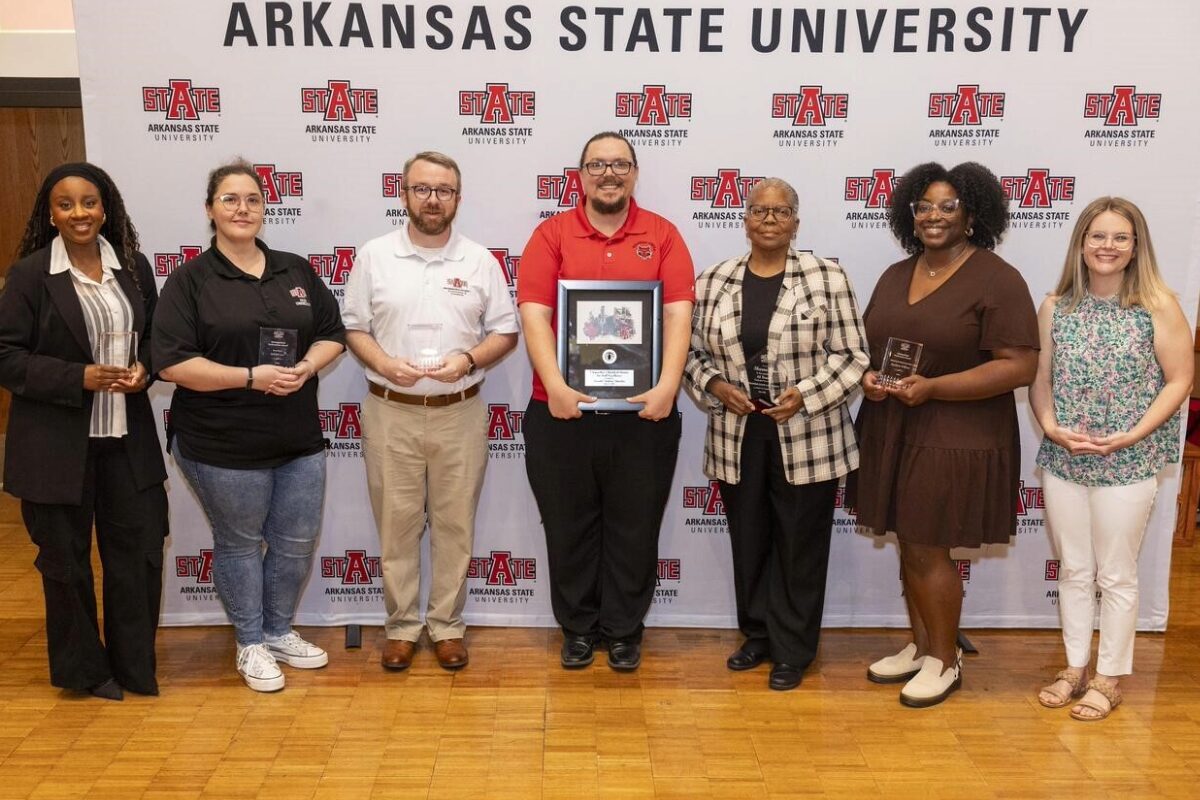 Group of seven people standing in front of an Arkansas State University backdrop, each holding an award or plaque at an event.