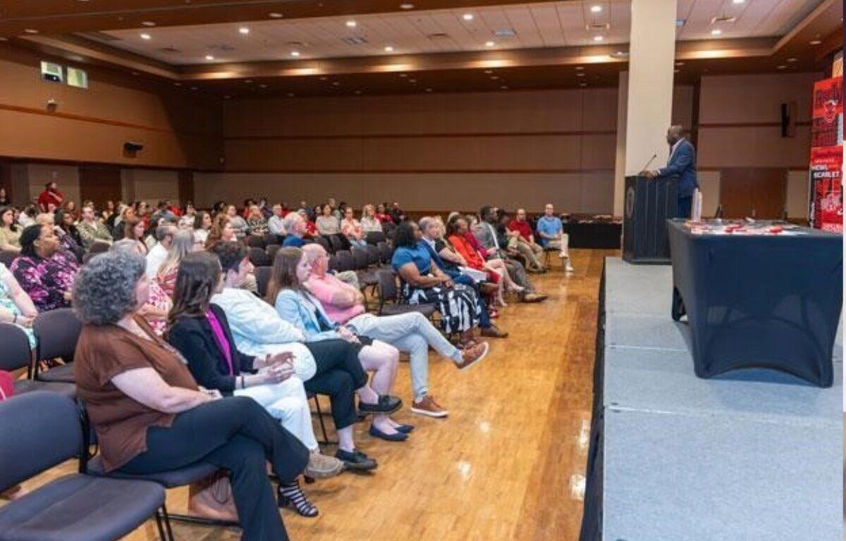 Speaker at a podium addressing a seated audience in a large hall for a conference or event.