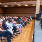Speaker at a podium addressing a seated audience in a large hall for a conference or event.