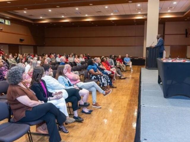 Speaker at a podium addressing a seated audience in a large hall for a conference or event.