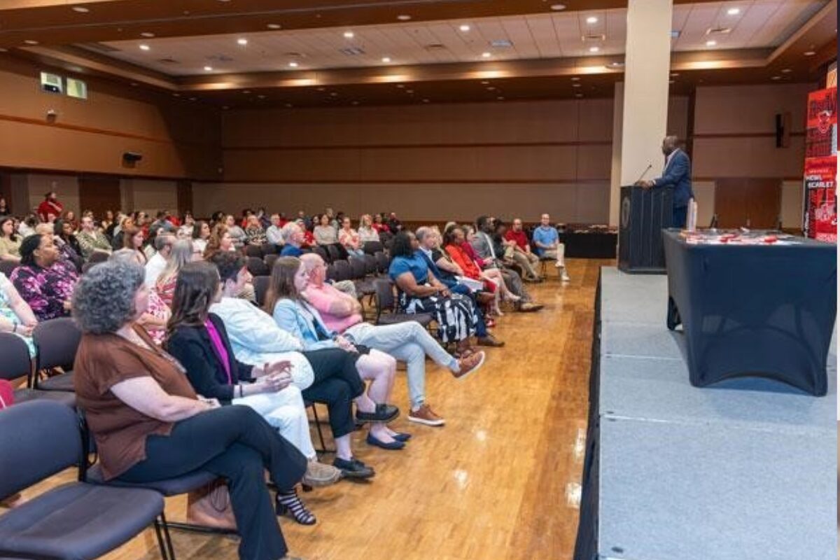 Speaker at a podium addressing a seated audience in a large hall for a conference or event.