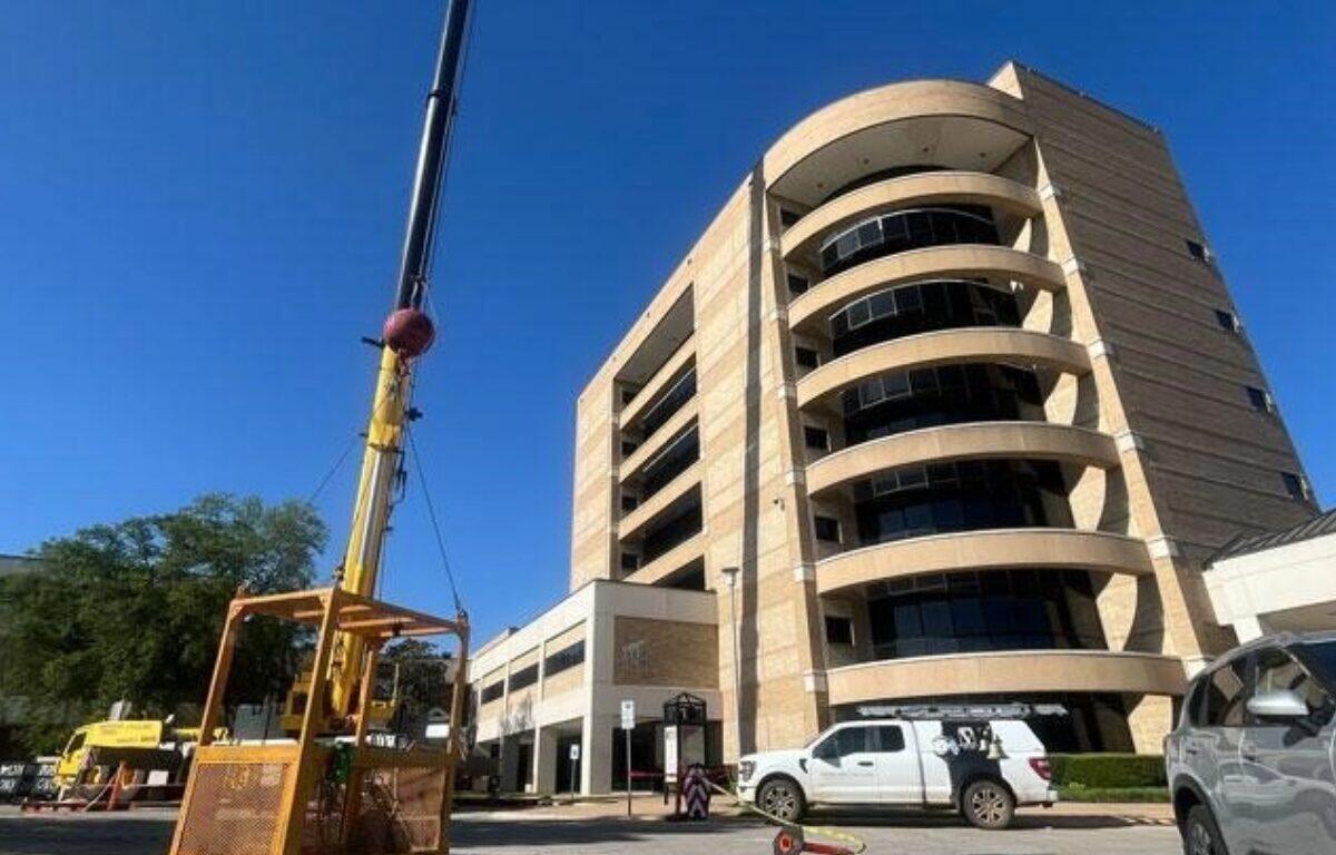 Construction crane beside a tall curved beige office building, with work vans parked in front.