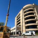 Construction crane beside a tall curved beige office building, with work vans parked in front.