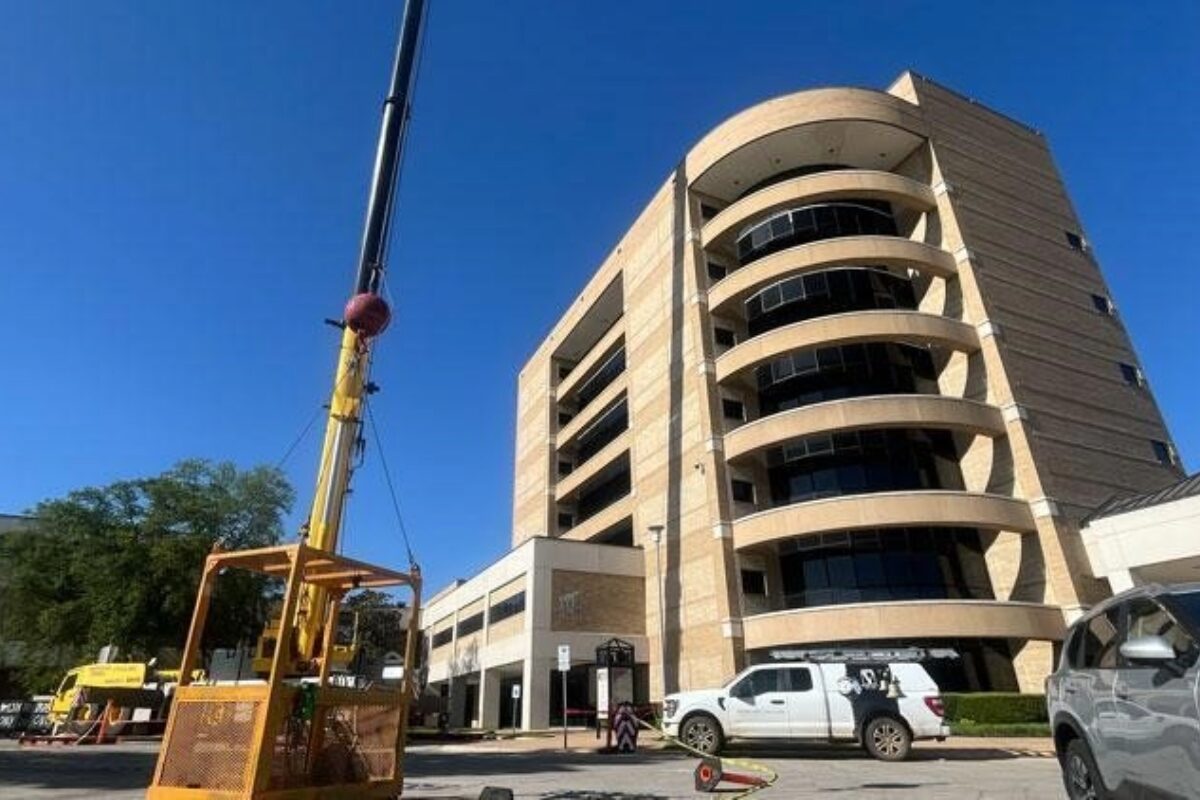 Construction crane beside a tall curved beige office building, with work vans parked in front.
