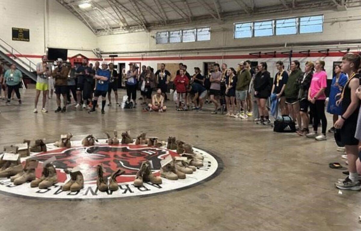 Group of people standing around a circular display of brown boots arranged on the gym floor, likely a memorial or tribute.