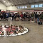 Group of people standing around a circular display of brown boots arranged on the gym floor, likely a memorial or tribute.