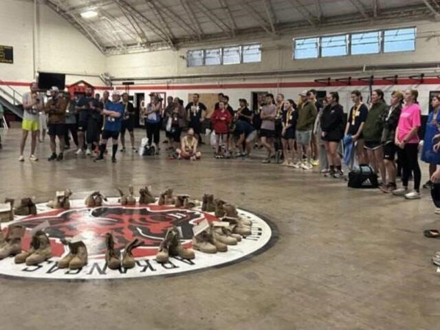 Group of people standing around a circular display of brown boots arranged on the gym floor, likely a memorial or tribute.
