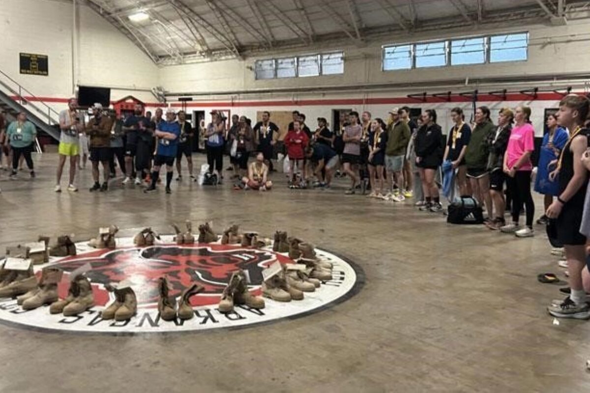 Group of people standing around a circular display of brown boots arranged on the gym floor, likely a memorial or tribute.