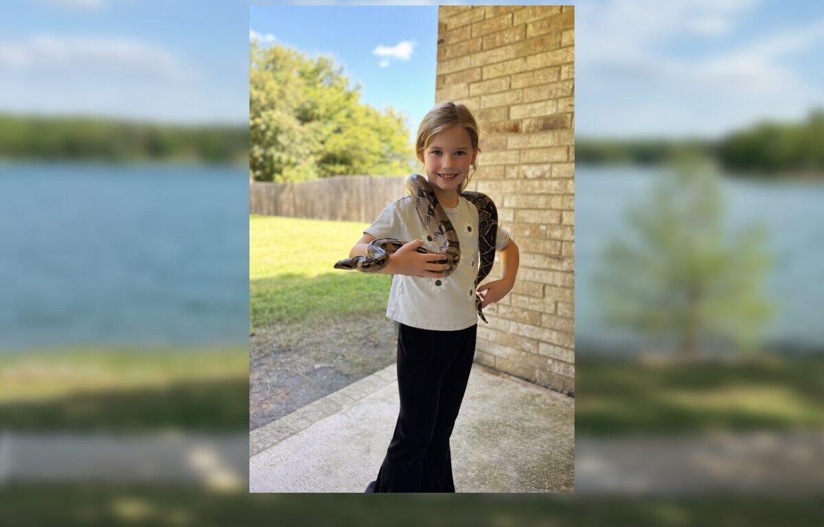 Smiling girl outdoors on a porch with a snake draped over her shoulders, holding its body with both hands.
