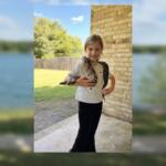 Smiling girl outdoors on a porch with a snake draped over her shoulders, holding its body with both hands.