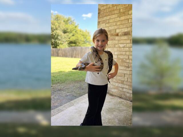 Smiling girl outdoors on a porch with a snake draped over her shoulders, holding its body with both hands.