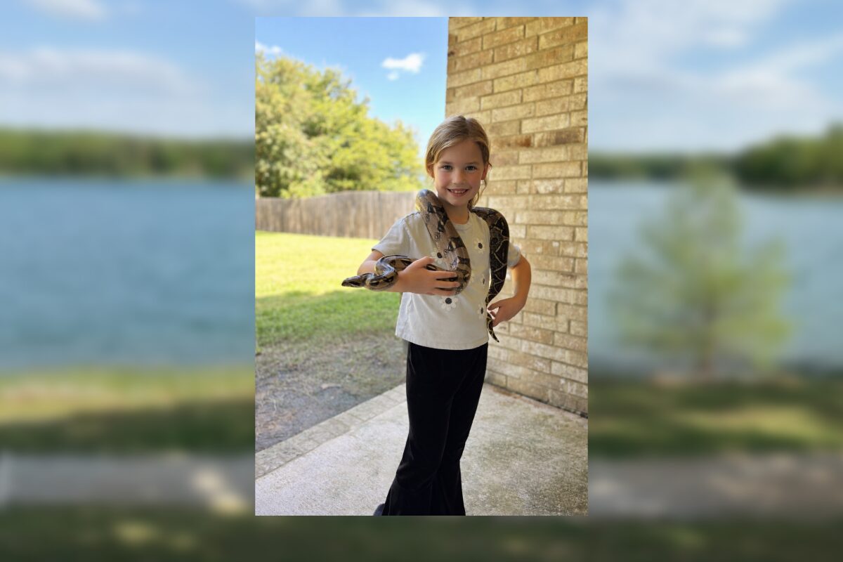 Smiling girl outdoors on a porch with a snake draped over her shoulders, holding its body with both hands.