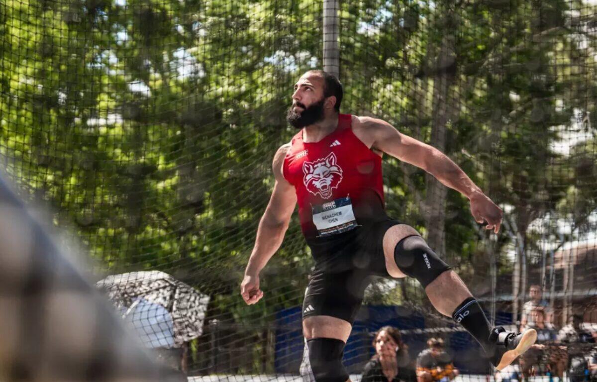 Male athlete in a red sleeveless shirt with a wolf logo mid-air in a jump, safety net and spectators behind him (outdoors)