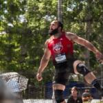 Male athlete in a red sleeveless shirt with a wolf logo mid-air in a jump, safety net and spectators behind him (outdoors)