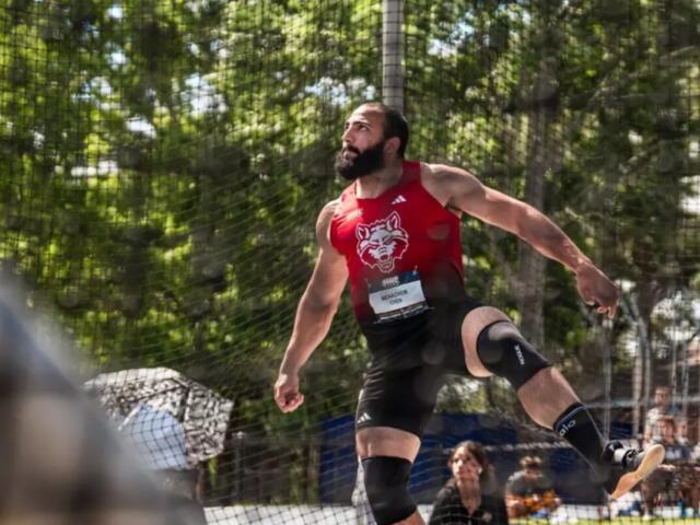Male athlete in a red sleeveless shirt with a wolf logo mid-air in a jump, safety net and spectators behind him (outdoors)