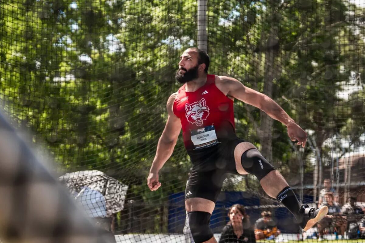 Male athlete in a red sleeveless shirt with a wolf logo mid-air in a jump, safety net and spectators behind him (outdoors)