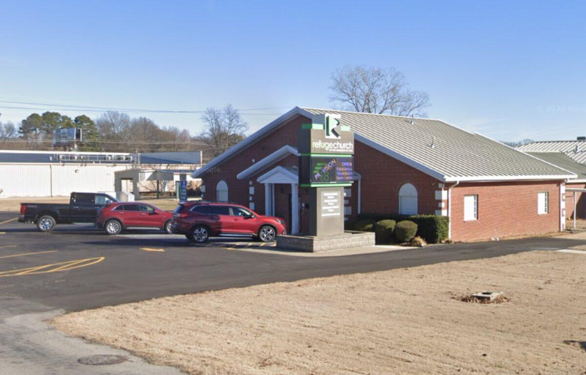 Brick church with a gabled roof and a sign for Refuge Church; cars parked in the front lot on a clear day.