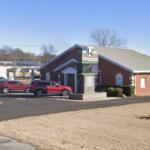 Brick church with a gabled roof and a sign for Refuge Church; cars parked in the front lot on a clear day.
