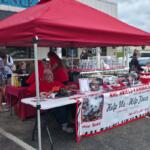 Outdoor pet adoption booth under a red canopy; people chat near cages with small dogs, banner reads 'Help Us – Help Them' for ARC Angels Animal Rescue.