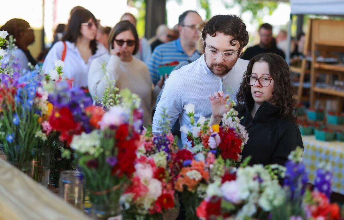 Couple stands at a flower stall, examining bright bouquets of red, pink, and purple flowers while other shoppers browse in the background.