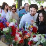 Couple stands at a flower stall, examining bright bouquets of red, pink, and purple flowers while other shoppers browse in the background.