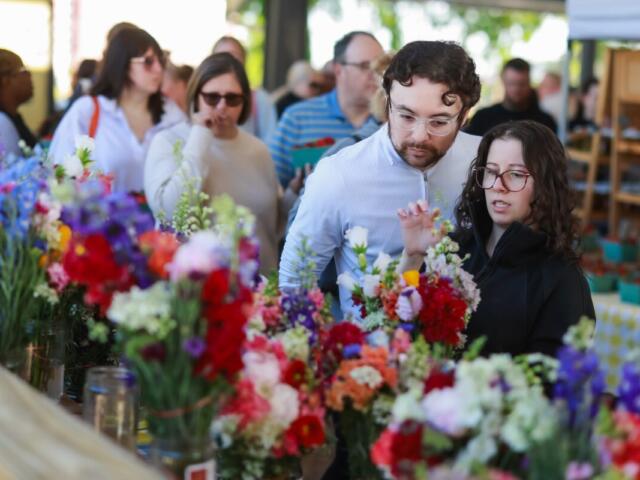 Couple stands at a flower stall, examining bright bouquets of red, pink, and purple flowers while other shoppers browse in the background.